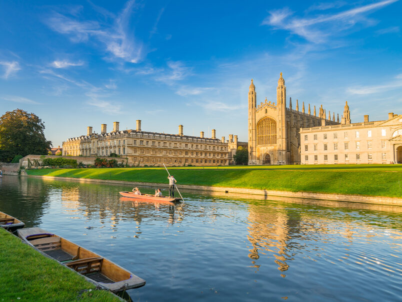 Blick auf das King’s College in Cambridge am Fluss Cam bei sonnigem Wetter; im Vordergrund liegen mehrere Boote am Ufer, auf dem Wasser fährt ein Punt mit Personen, und das historische Gebäude spiegelt sich im Fluss.
