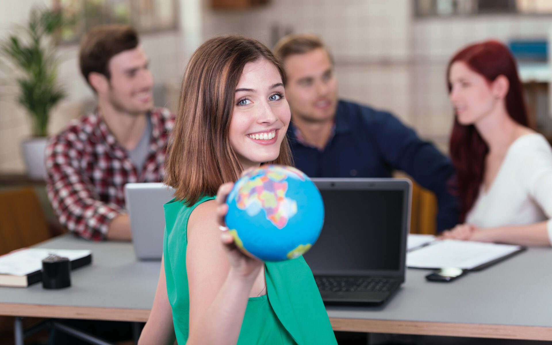 Lächelnde junge Frau hält einen kleinen Globus in die Kamera; im Hintergrund sitzen drei weitere Personen an einem Tisch mit Laptops und Unterlagen.