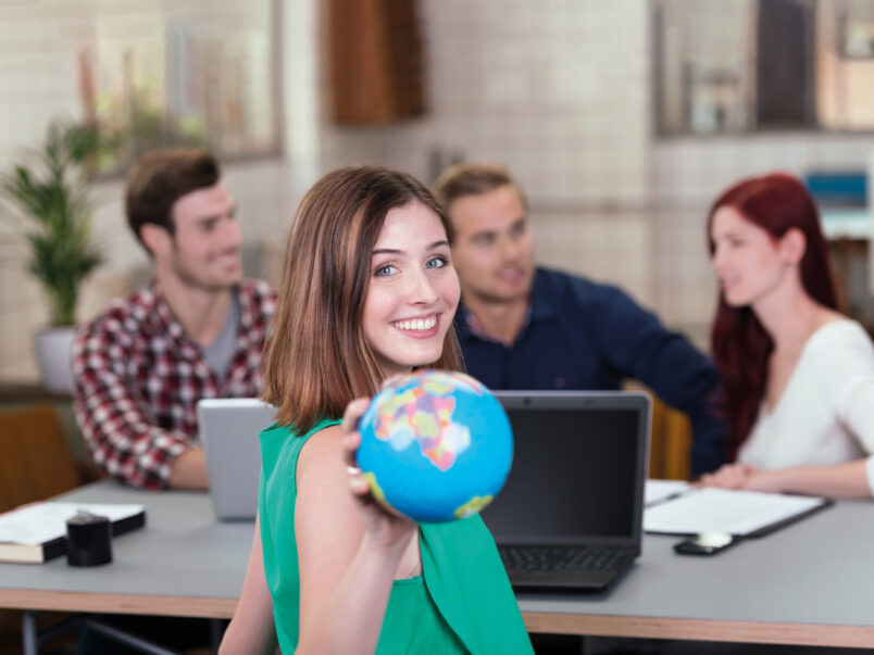 Lächelnde junge Frau hält einen kleinen Globus in die Kamera; im Hintergrund sitzen drei weitere Personen an einem Tisch mit Laptops und Unterlagen.