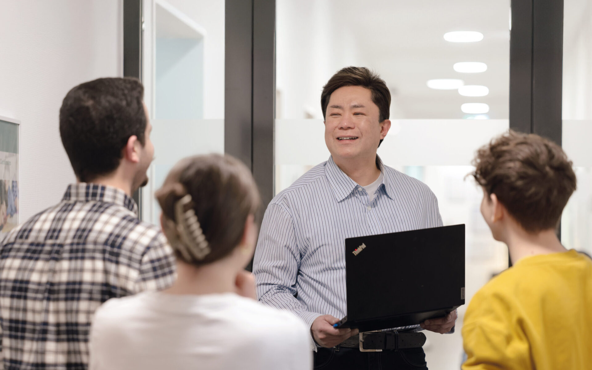 Ein Mann mit Laptop spricht in einem hellen Flur zu einer kleinen Gruppe von Personen, die ihm zugewandt stehen; die Szene wirkt wie eine Schulung oder kurze Präsentation.
