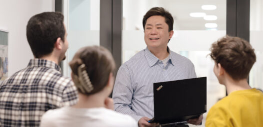 Ein Mann mit Laptop spricht in einem hellen Flur zu einer kleinen Gruppe von Personen, die ihm zugewandt stehen; die Szene wirkt wie eine Schulung oder kurze Präsentation.