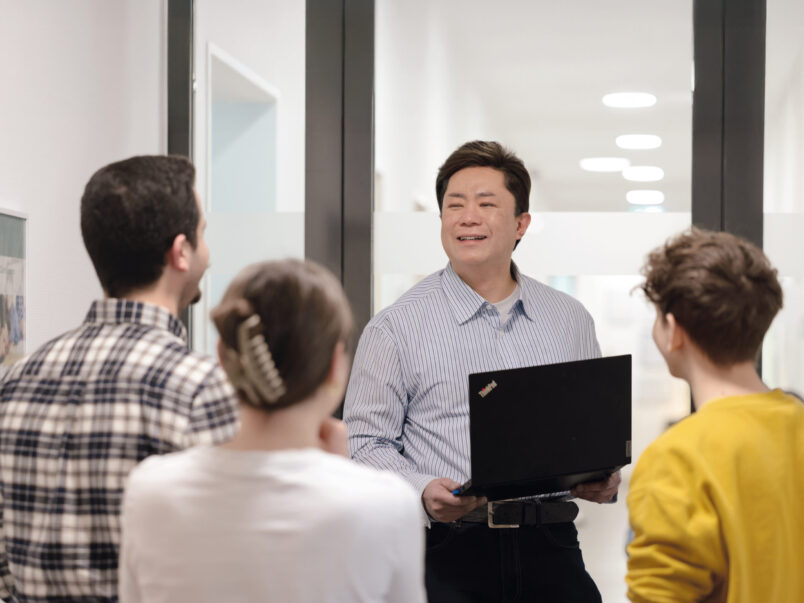 Ein Mann mit Laptop spricht in einem hellen Flur zu einer kleinen Gruppe von Personen, die ihm zugewandt stehen; die Szene wirkt wie eine Schulung oder kurze Präsentation.