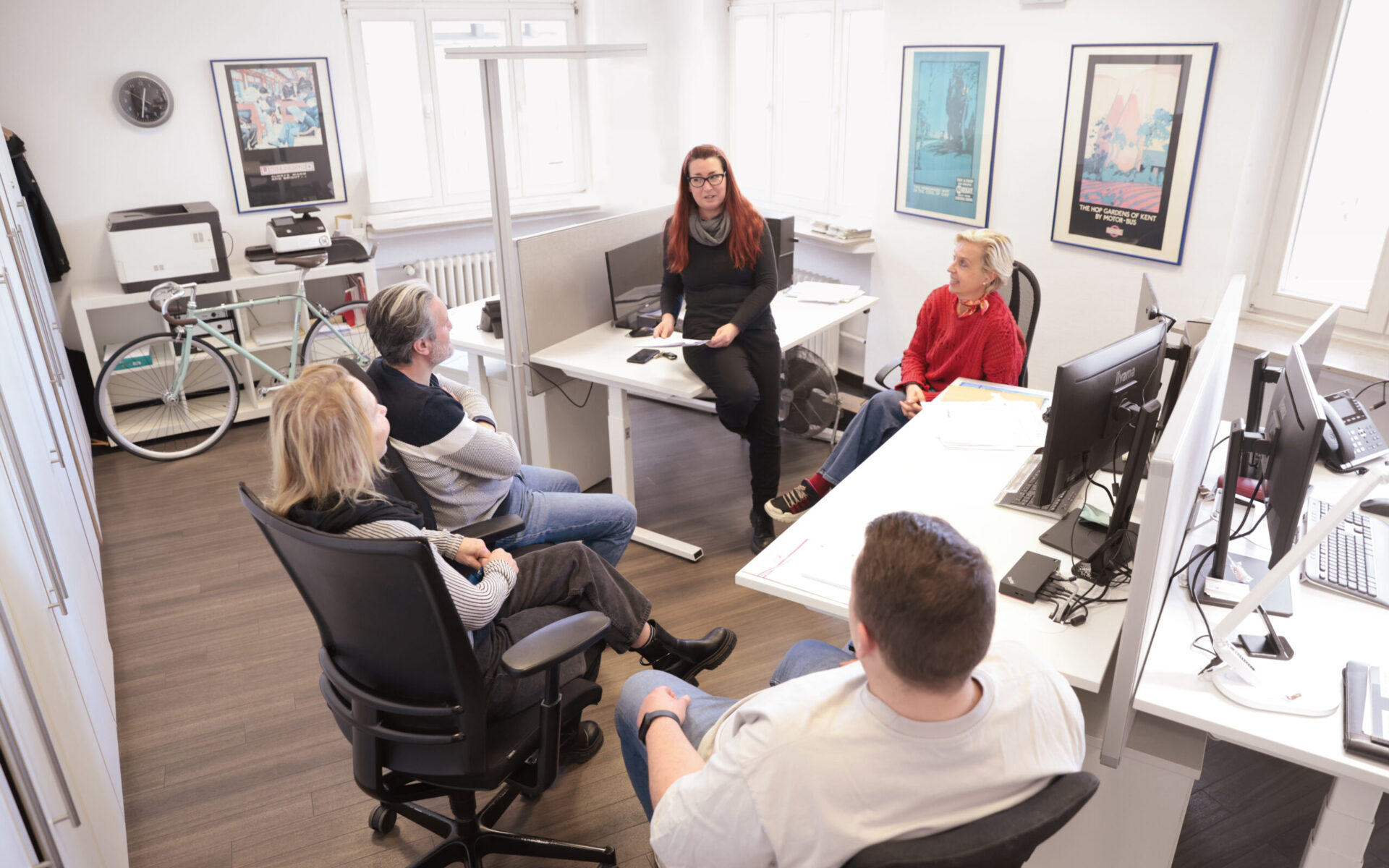 Fünf Personen sitzen in einem hellen Büro zu einer Besprechung zusammen; eine Frau spricht vorne im Raum, während die anderen auf Bürostühlen zuhören. Schreibtische mit Monitoren und Arbeitsplätzen umgeben die Gruppe.
