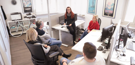 Fünf Personen sitzen in einem hellen Büro zu einer Besprechung zusammen; eine Frau spricht vorne im Raum, während die anderen auf Bürostühlen zuhören. Schreibtische mit Monitoren und Arbeitsplätzen umgeben die Gruppe.