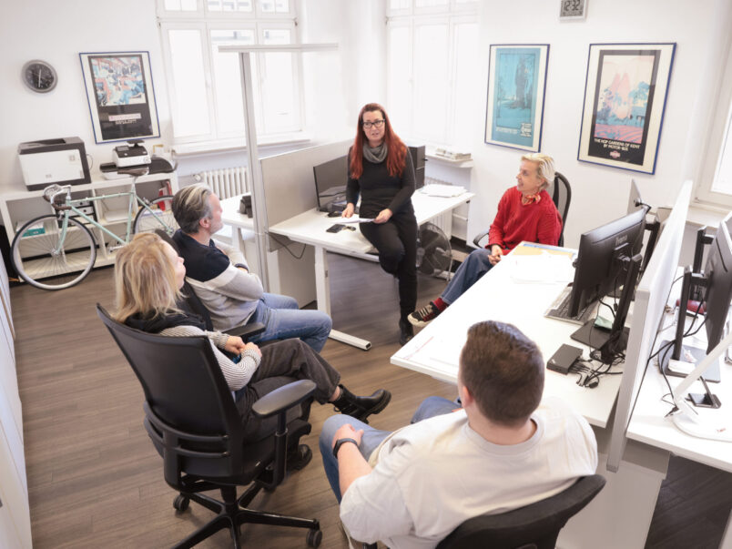 Fünf Personen sitzen in einem hellen Büro zu einer Besprechung zusammen; eine Frau spricht vorne im Raum, während die anderen auf Bürostühlen zuhören. Schreibtische mit Monitoren und Arbeitsplätzen umgeben die Gruppe.