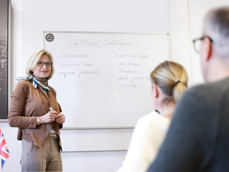 Dozentin steht vor einem Whiteboard und spricht zu einer kleinen Gruppe; auf der Tafel stehen Stichpunkte zu künstlicher Intelligenz mit Vor- und Nachteilen.