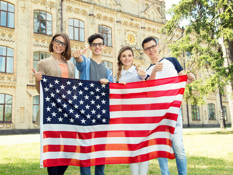 Vier junge Erwachsene stehen vor einem historischen Gebäude im Freien, halten eine große US-Flagge und zeigen mit Daumen nach oben in die Kamera.