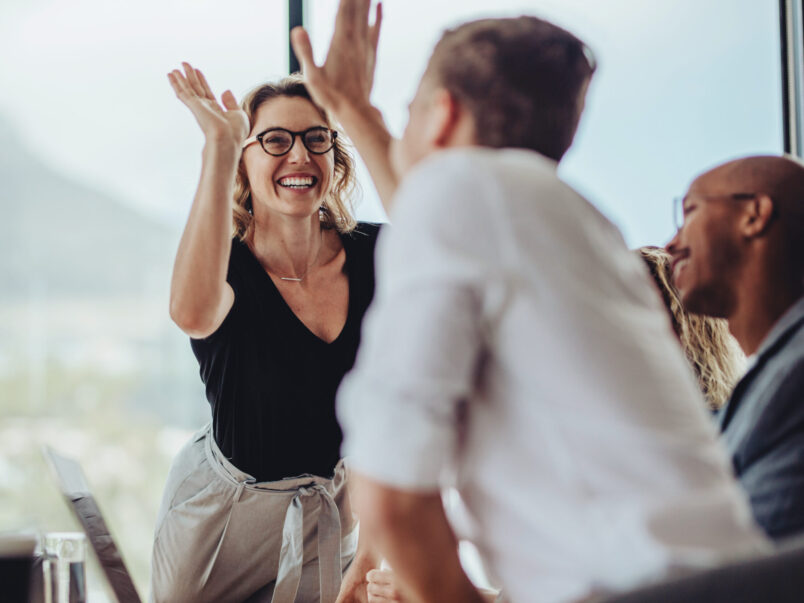 Lächelnde Frau in einem Besprechungsraum gibt einem Kollegen ein High Five; weitere Personen sitzen im Hintergrund, die Szene wirkt wie ein gemeinsamer Erfolg im Team.