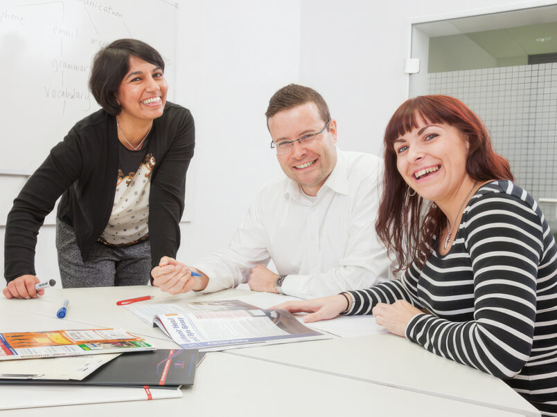 Drei lächelnde Personen sitzen gemeinsam an einem Besprechungstisch mit Unterlagen und Stiften; im Hintergrund ist ein Whiteboard zu sehen, die Szene wirkt wie ein Sprachkurs oder Teammeeting.