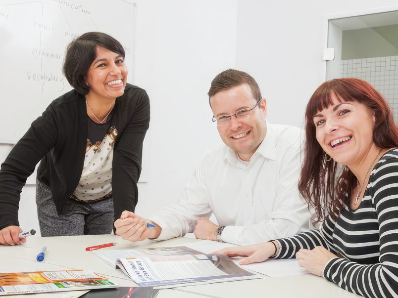 Drei lächelnde Personen sitzen gemeinsam an einem Besprechungstisch mit Unterlagen und Stiften; im Hintergrund ist ein Whiteboard zu sehen, die Szene wirkt wie ein Sprachkurs oder Teammeeting.