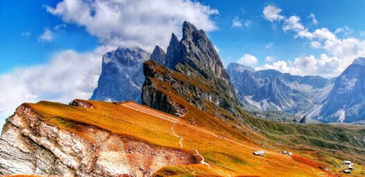 Alpine Berglandschaft mit schroffen Felsgipfeln unter blauem Himmel und großen Wolken; im Vordergrund goldbraune Wiesen, ein geschwungener Wanderweg und einige kleine Hütten.
