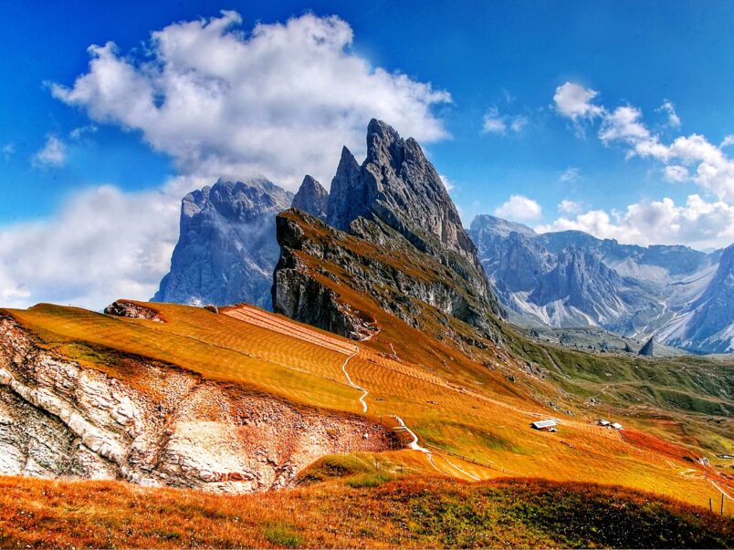 Alpine Berglandschaft mit schroffen Felsgipfeln unter blauem Himmel und großen Wolken; im Vordergrund goldbraune Wiesen, ein geschwungener Wanderweg und einige kleine Hütten.