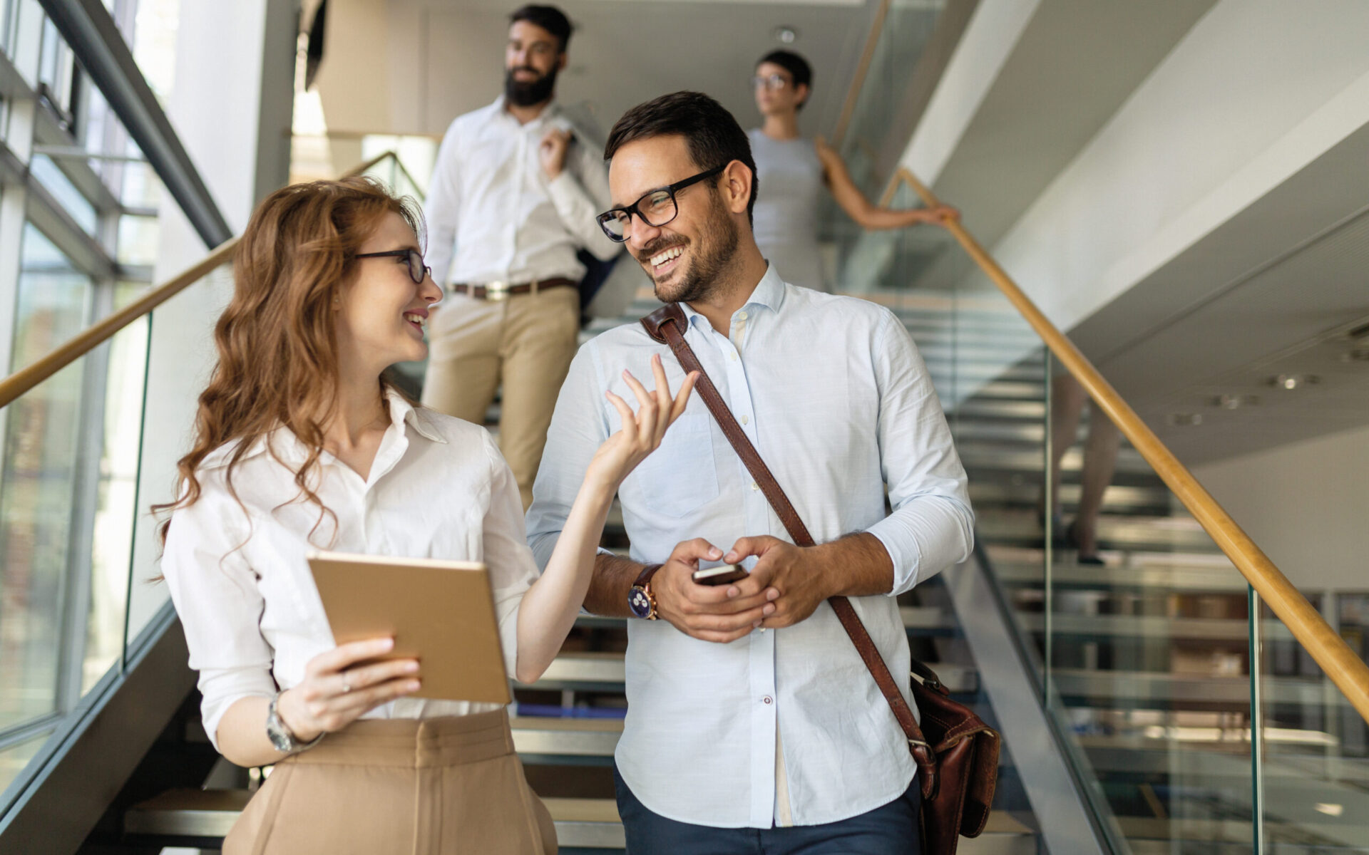 Zwei Personen gehen gemeinsam eine Treppe in einem modernen Gebäude hinunter und unterhalten sich; die Frau hält ein Tablet, der Mann ein Smartphone. Im Hintergrund folgen zwei weitere Personen.