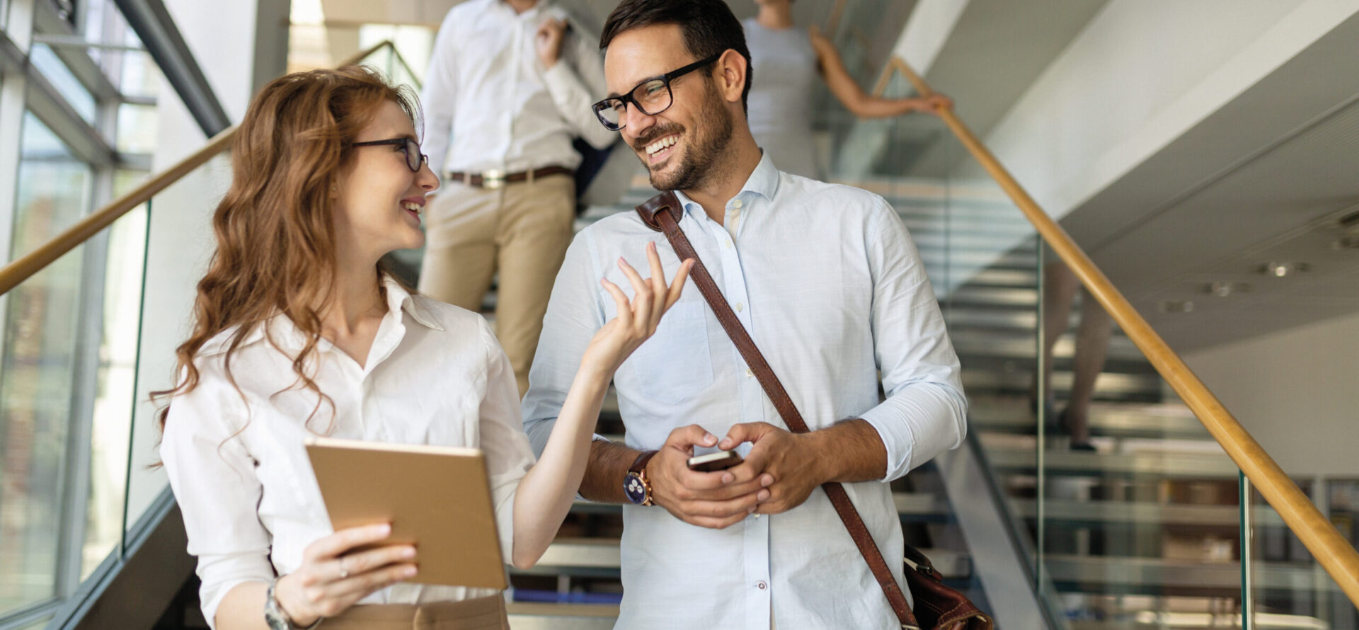 Zwei Personen gehen gemeinsam eine Treppe in einem modernen Gebäude hinunter und unterhalten sich; die Frau hält ein Tablet, der Mann ein Smartphone. Im Hintergrund folgen zwei weitere Personen.