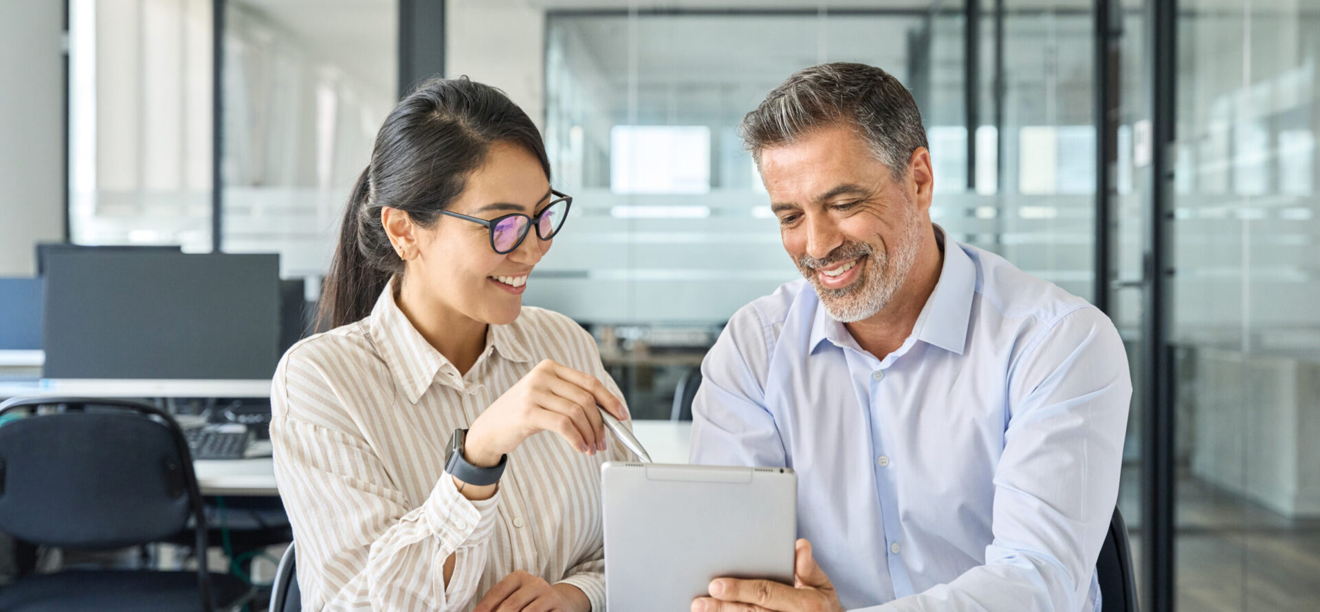 Zwei Personen sitzen in einem modernen Büro an einem Tisch und schauen gemeinsam auf ein Tablet; die Frau zeigt mit einem Stift auf den Bildschirm, während der Mann das Gerät hält.