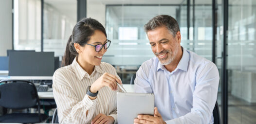 Zwei Personen sitzen in einem modernen Büro an einem Tisch und schauen gemeinsam auf ein Tablet; die Frau zeigt mit einem Stift auf den Bildschirm, während der Mann das Gerät hält.