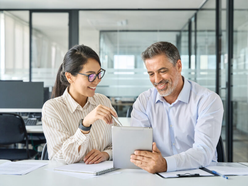 Zwei Personen sitzen in einem modernen Büro an einem Tisch und schauen gemeinsam auf ein Tablet; die Frau zeigt mit einem Stift auf den Bildschirm, während der Mann das Gerät hält.