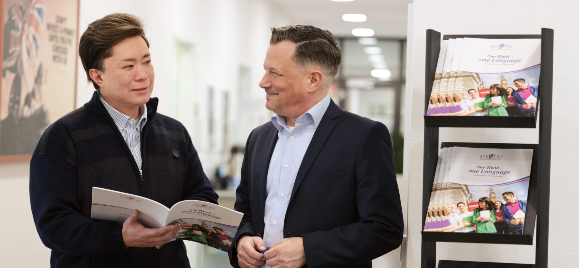 Zwei Männer stehen in einem Flur und unterhalten sich über eine Broschüre; daneben befindet sich ein Prospektständer mit mehreren Informationsheften von STEVENS.