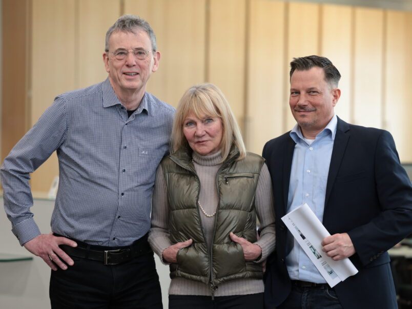 Drei Erwachsene stehen nebeneinander in einem Innenraum und blicken in die Kamera; der Mann rechts hält einige Unterlagen in der Hand.