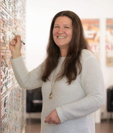 Lächelnde Frau steht vor einem großen Wandplan mit vielen farbigen Einträgen und markiert etwas mit einem Stift.