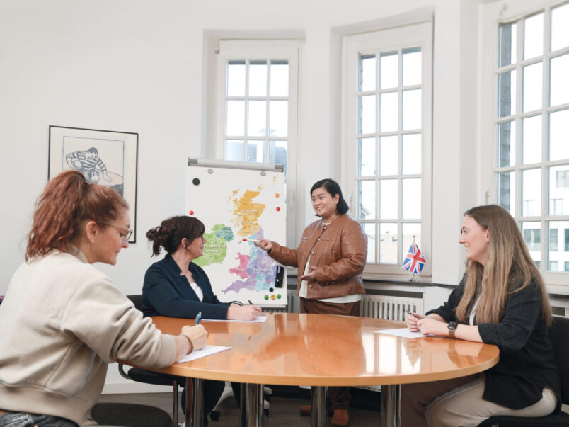Kleine Unterrichts- oder Besprechungsrunde in einem hellen Raum: Eine Frau steht vor einer Karte der britischen Inseln und erklärt etwas, während drei weitere Frauen am runden Tisch zuhören und Notizen machen.