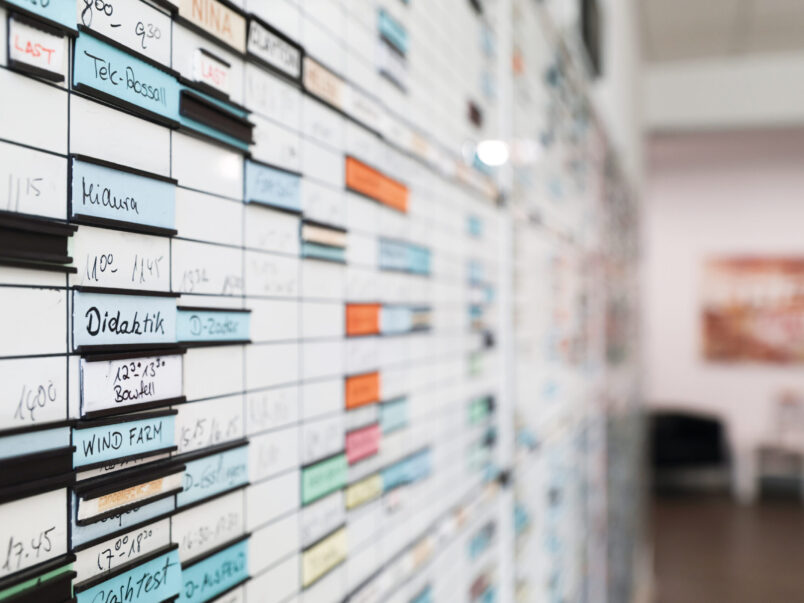 Nahaufnahme eines Wandplans oder Stundenplans mit beschrifteten, farbigen Einsteckstreifen; im Hintergrund ist ein Flur oder Raum unscharf zu sehen.