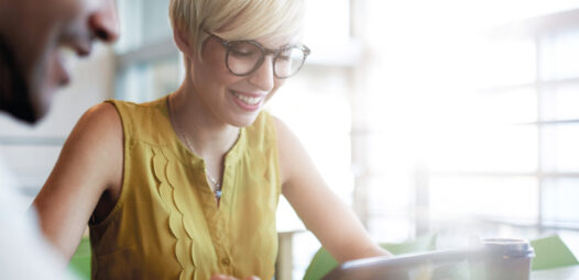 Lächelnde Frau mit Kurzhaarfrisur und Brille sitzt an einem Tisch und schaut auf ein Tablet; im Vordergrund ist unscharf eine weitere Person zu sehen, daneben steht ein Kaffeebecher.
