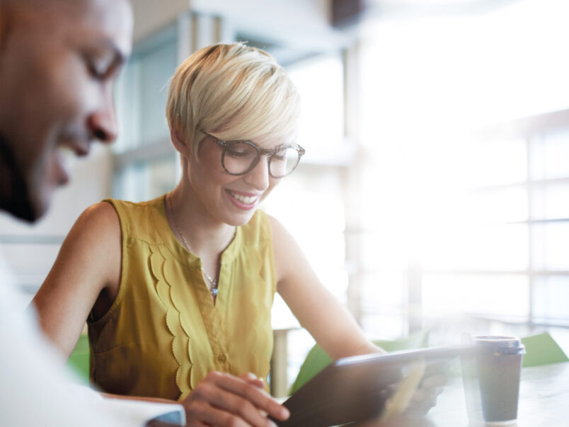 Lächelnde Frau mit Kurzhaarfrisur und Brille sitzt an einem Tisch und schaut auf ein Tablet; im Vordergrund ist unscharf eine weitere Person zu sehen, daneben steht ein Kaffeebecher.