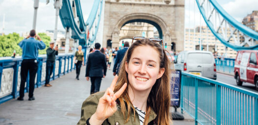 Junge Frau steht auf der Tower Bridge in London, lächelt in die Kamera und zeigt ein Peace-Zeichen; im Hintergrund sind der Brückenturm, Fußgänger und Fahrzeuge zu sehen.