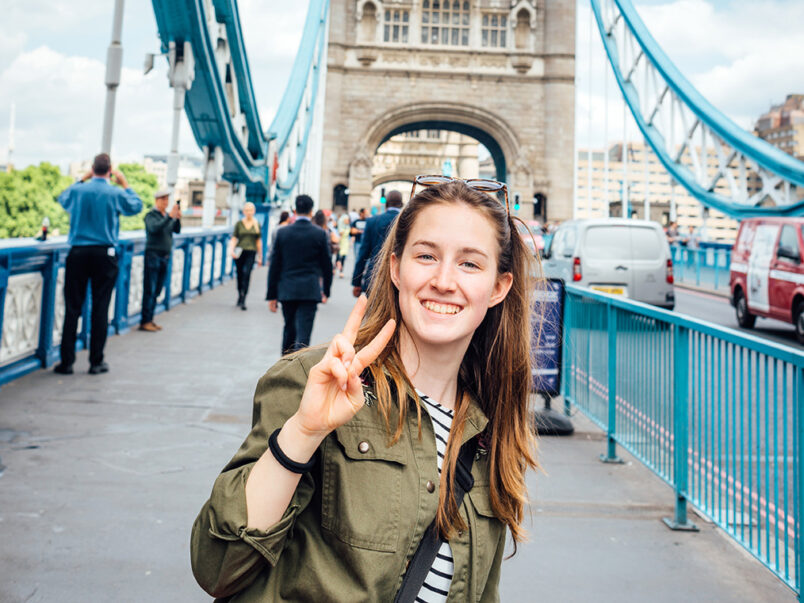 Junge Frau steht auf der Tower Bridge in London, lächelt in die Kamera und zeigt ein Peace-Zeichen; im Hintergrund sind der Brückenturm, Fußgänger und Fahrzeuge zu sehen.