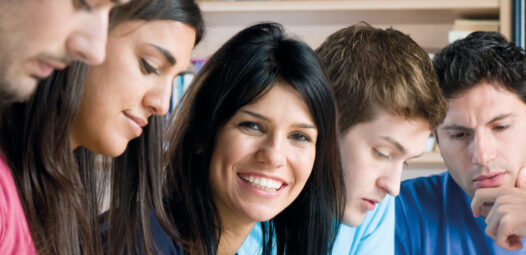 Fünf junge Erwachsene sitzen gemeinsam an einem Tisch in einer Bibliothek oder einem Lernraum, schreiben Notizen und arbeiten zusammen; eine Frau in der Mitte lächelt in die Kamera.