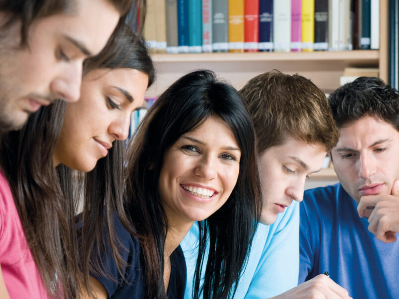 Fünf junge Erwachsene sitzen gemeinsam an einem Tisch in einer Bibliothek oder einem Lernraum, schreiben Notizen und arbeiten zusammen; eine Frau in der Mitte lächelt in die Kamera.