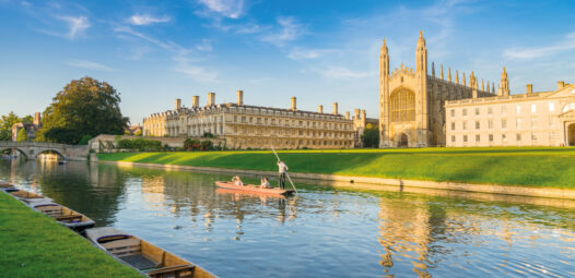 Blick auf das King’s College in Cambridge am Fluss Cam bei sonnigem Wetter; im Vordergrund liegen mehrere Boote am Ufer, auf dem Wasser fährt ein Punt mit Personen, und das historische Gebäude spiegelt sich im Fluss.
