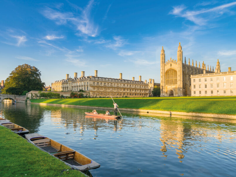 Blick auf das King’s College in Cambridge am Fluss Cam bei sonnigem Wetter; im Vordergrund liegen mehrere Boote am Ufer, auf dem Wasser fährt ein Punt mit Personen, und das historische Gebäude spiegelt sich im Fluss.