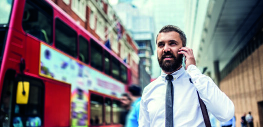 Ein Geschäftsmann telefoniert auf einer belebten Straße in der Stadt; neben ihm fährt ein roter Doppeldeckerbus vorbei.