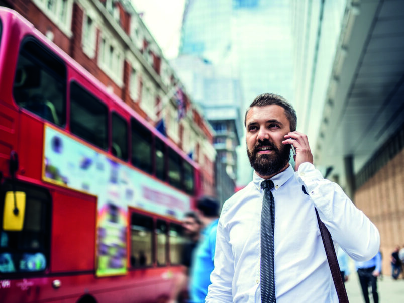 Ein Geschäftsmann telefoniert auf einer belebten Straße in der Stadt; neben ihm fährt ein roter Doppeldeckerbus vorbei.