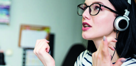 Frau mit Brille und Kopfhörern spricht aufmerksam und gestikuliert mit den Händen; die Szene wirkt wie ein Online-Gespräch oder Sprachtraining.