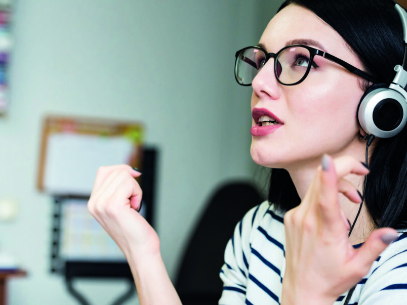 Frau mit Brille und Kopfhörern spricht aufmerksam und gestikuliert mit den Händen; die Szene wirkt wie ein Online-Gespräch oder Sprachtraining.