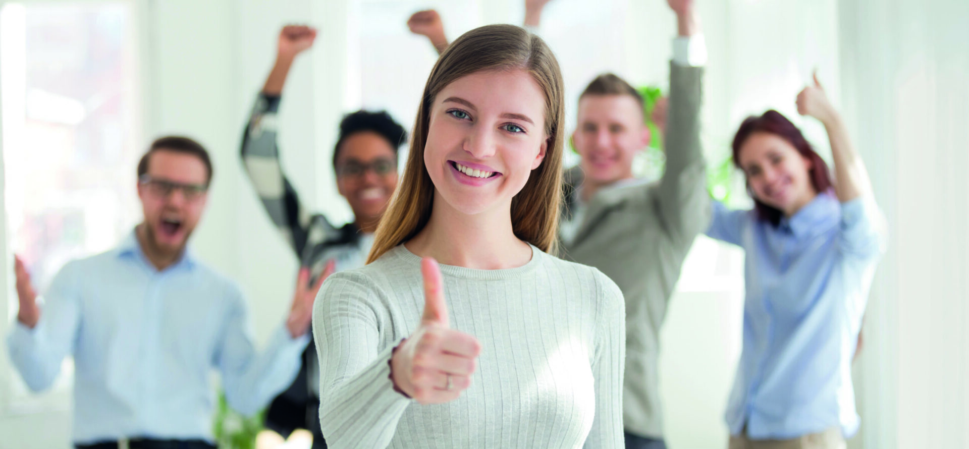 Lächelnde junge Frau steht im Vordergrund und zeigt den Daumen nach oben; im Hintergrund jubeln vier weitere Personen in einem hellen Raum.