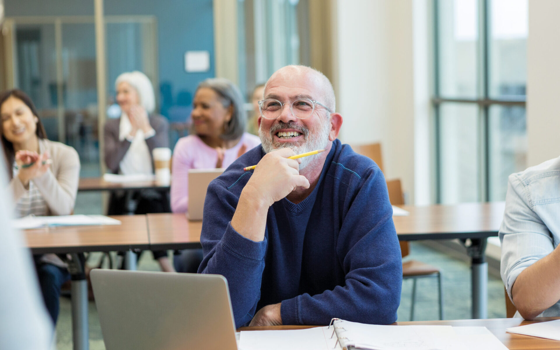 Lächelnder älterer Mann mit Brille sitzt in einem Seminarraum an einem Tisch mit Laptop und Unterlagen; er hält einen Stift in der Hand und hört aufmerksam zu. Im Hintergrund sitzen weitere Teilnehmende.