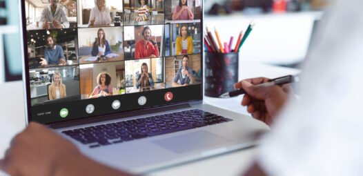 Auf dem Bildschirm eines Laptops ist eine Videokonferenz mit mehreren Teilnehmenden in Kachelansicht zu sehen; im Vordergrund hält eine Person einen Stift in der Hand. Daneben steht ein Becher mit bunten Stiften.