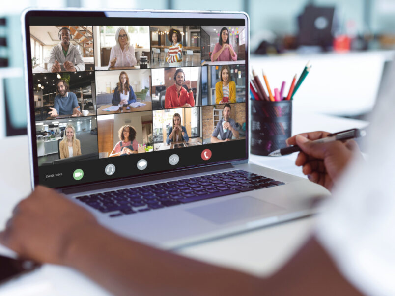 Auf dem Bildschirm eines Laptops ist eine Videokonferenz mit mehreren Teilnehmenden in Kachelansicht zu sehen; im Vordergrund hält eine Person einen Stift in der Hand. Daneben steht ein Becher mit bunten Stiften.