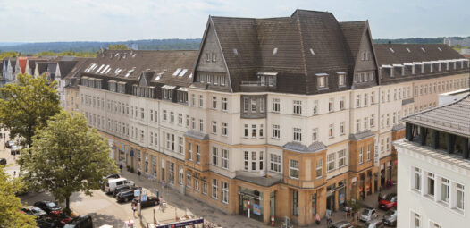 Luftige Stadtansicht mit einem großen Eckgebäude mit dunklem Dach an einer Straßenkreuzung; unten sind parkende Autos, Bäume und ein Zugang zu einer U-Bahn-Station zu sehen.