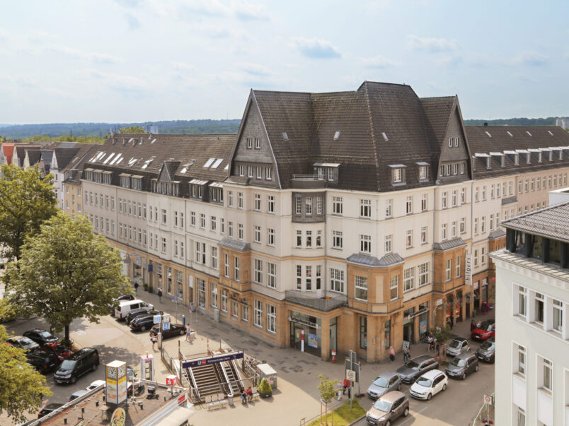 Luftige Stadtansicht mit einem großen Eckgebäude mit dunklem Dach an einer Straßenkreuzung; unten sind parkende Autos, Bäume und ein Zugang zu einer U-Bahn-Station zu sehen.