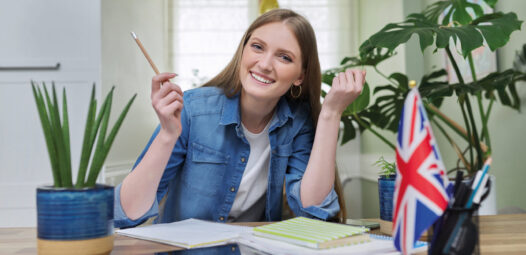 Lächelnde junge Frau sitzt an einem Tisch mit Heften und Stift; daneben stehen Zimmerpflanzen und eine kleine britische Flagge. Die Szene wirkt wie Lernen oder Englischunterricht zu Hause.