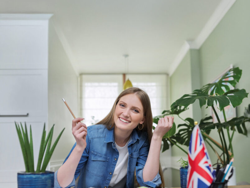 Lächelnde junge Frau sitzt an einem Tisch mit Heften und Stift; daneben stehen Zimmerpflanzen und eine kleine britische Flagge. Die Szene wirkt wie Lernen oder Englischunterricht zu Hause.