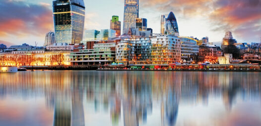 Skyline von London bei Sonnenuntergang mit modernen Hochhäusern am Wasser; die Gebäude und farbigen Wolken spiegeln sich ruhig in der Themse.
