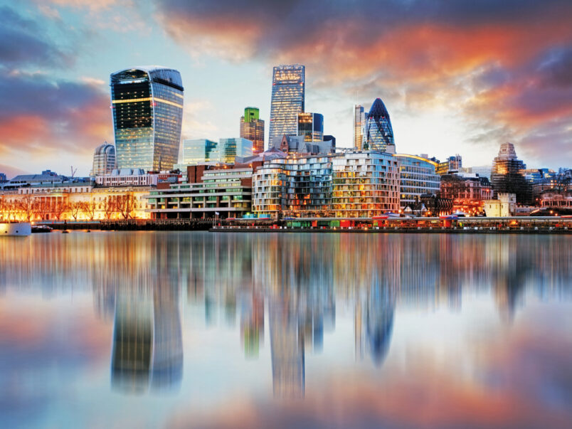 Skyline von London bei Sonnenuntergang mit modernen Hochhäusern am Wasser; die Gebäude und farbigen Wolken spiegeln sich ruhig in der Themse.