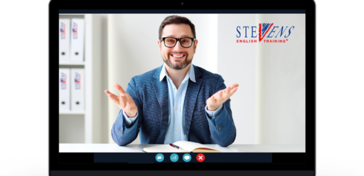 Auf dem Bildschirm eines Laptops ist ein lächelnder Mann in einem Videoanruf zu sehen; er sitzt an einem Schreibtisch mit offenem Buch und gestikuliert mit beiden Händen. Oben rechts im Bild steht das Logo von STEVENS English Training.