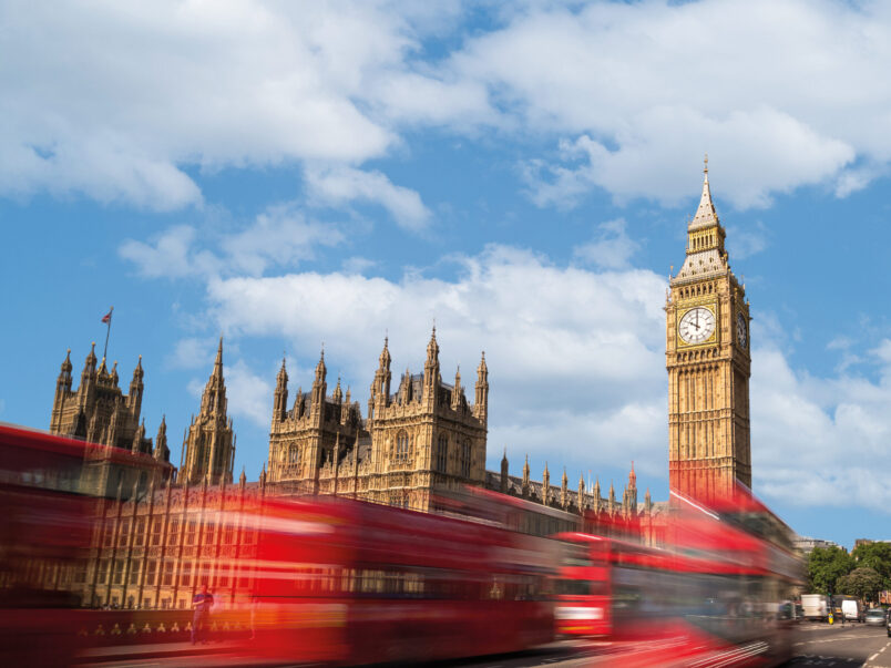 Blick auf den Elizabeth Tower und das Parlamentsgebäude in London unter blauem Himmel; im Vordergrund fahren rote Doppeldeckerbusse mit Bewegungsunschärfe über die Straße.